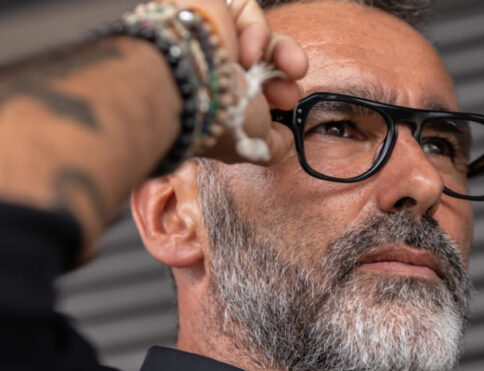 Publicité pour les lunettes MSW : un homme élégant aux cheveux gris, à la barbe et à la moustache grises, avec des tatouages et des bracelets, tient une paire de lunettes optique noires en acétate de la marque MSW