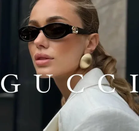 Femme élégante portant des lunettes de soleil Gucci noires avec le logo doré sur la branche, cheveux attachés en arrière, vêtue d'une veste blanche et de boucles d'oreilles dorées.